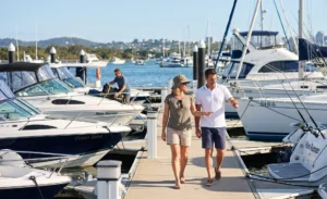 A couple walking along a sunlit marina dock, looking intently at various powerboats and sailboats moored in an Australian setting.